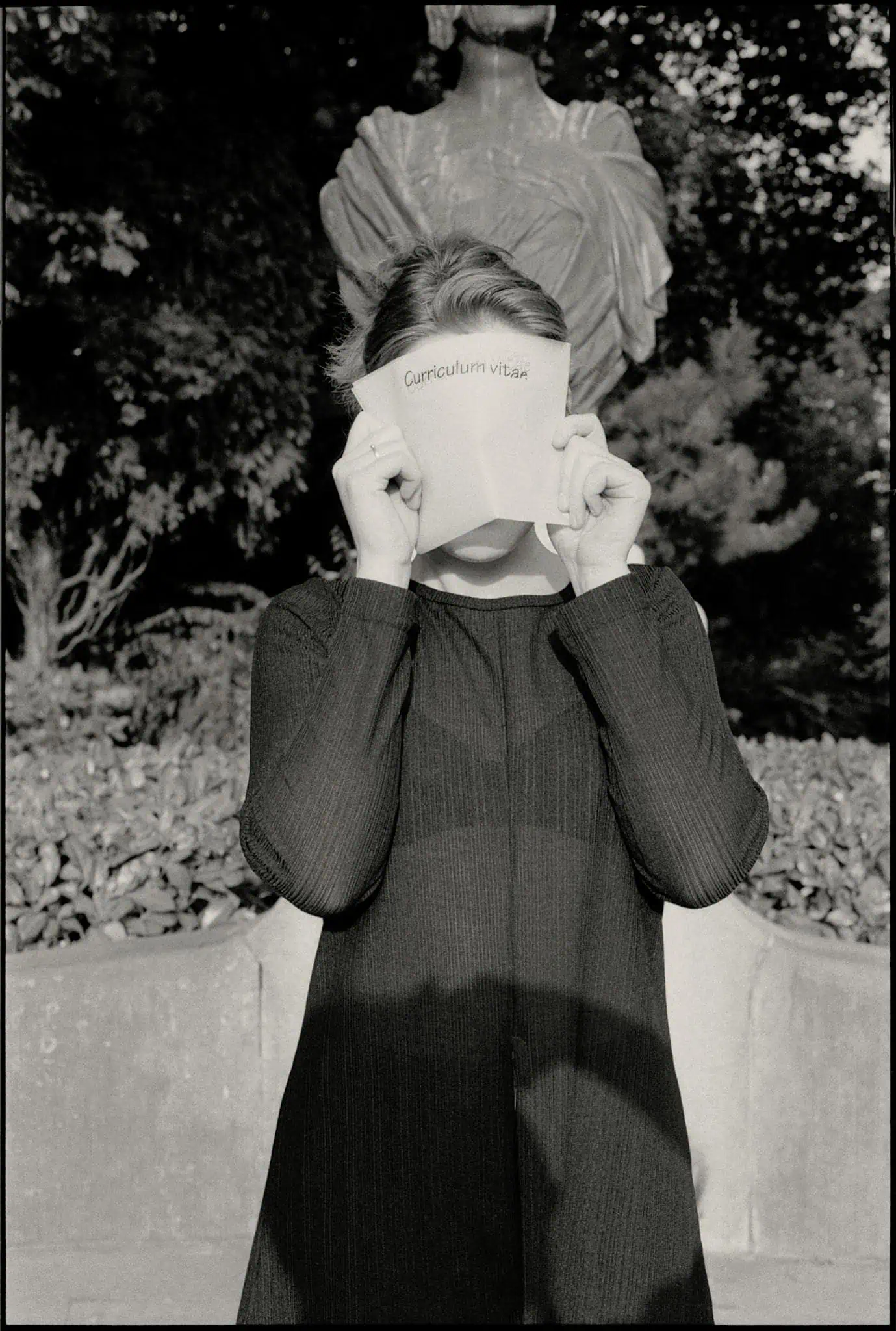 Black and white photo of a woman covering face with CV, standing outside.